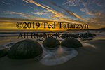 A dramatic golden and blue sunrise with the Moeraki boulders foreground splashed with time lapsed waves.