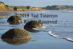 Three Moeraki boulders are lapped by wave foam a the shore with a long view of the coastline.