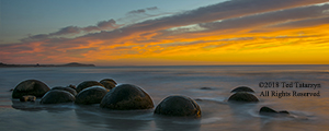 A dramatic yellow and orange sunrise panoramic image of a line of Moeraki boulders with waves lapping at the bottom of the boulders.